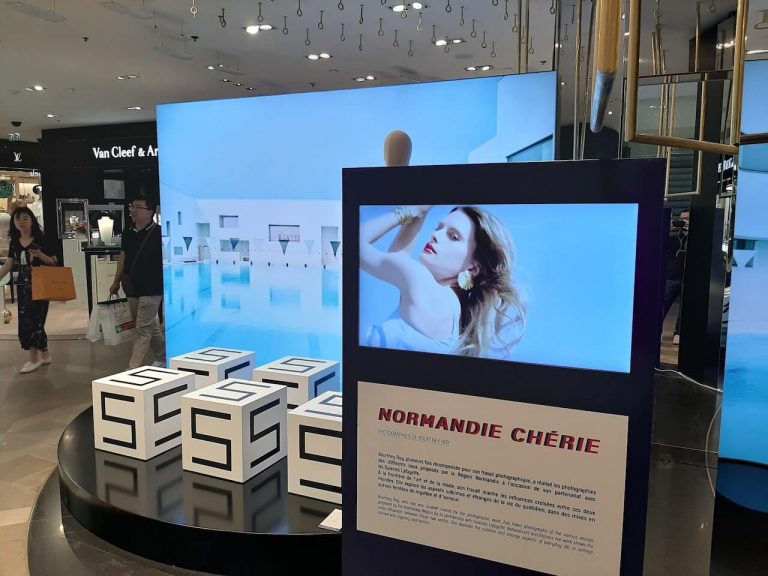 Totem noir avec écran diffusant une exposition photo, devant un grand mur d'images dans un grand magasin