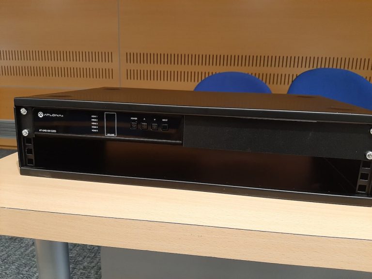 Commutateur HDMI Atlona monté dans un rack posé sur une table, panneaux acoustiques en bois en arrière-plan