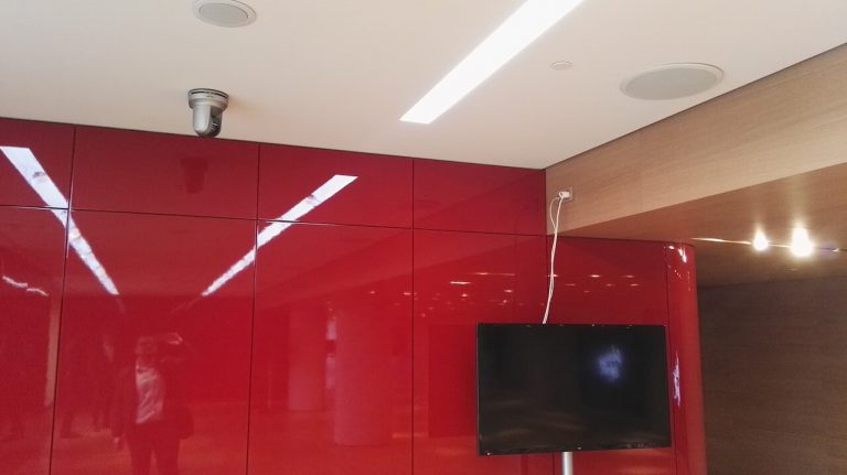 Caméra motorisée au plafond, enceintes encastrées et écran mural dans un espace d'accueil au mur rouge laqué