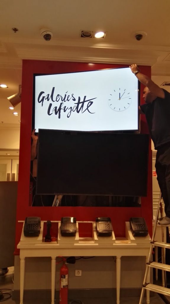 Technicien sur escabeau installant deux écrans superposés dans une niche murale rouge, logo Galeries Lafayette affiché