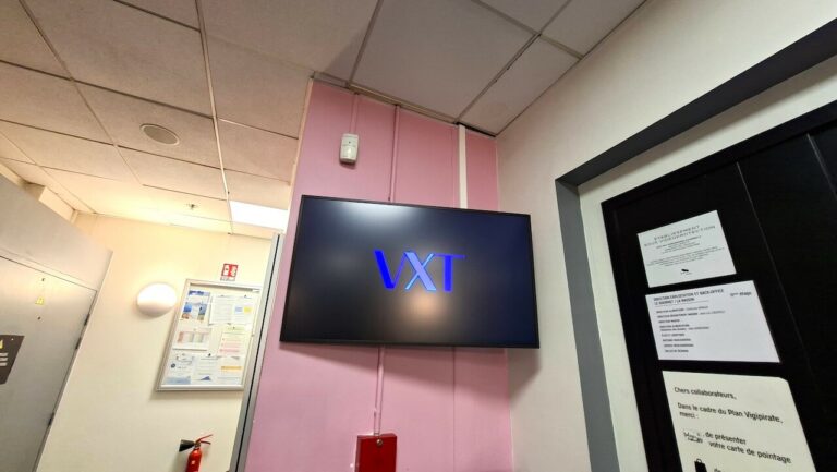 Écran affichant le logo VXT dans un espace réservé au personnel, à côté de panneaux de consignes internes